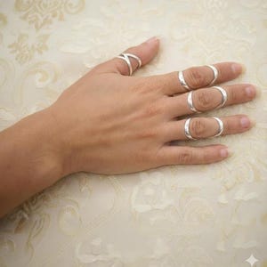 May include: A hand wearing several silver rings. The rings include a criss-cross design on the thumb and oval-shaped rings on the other fingers. The rings are simple and elegant, with a polished finish.