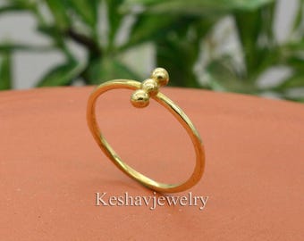 May include: A delicate gold ring with three small, round beads clustered together. The ring band is thin and smooth, and the beads are evenly spaced. The ring is set against a blurred green and brown background, with the text "Keshavjewelry" at the bottom.