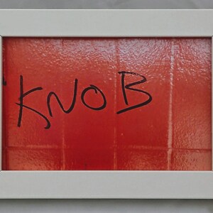 May include: A white framed picture with a red background and the word "KNOB" written in black marker.