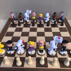 May include: A Peanuts-themed chess set with white and brown wooden board. The chess pieces are characters from the Peanuts comic strip, including Snoopy, Charlie Brown, Lucy, and Linus. The pieces are arranged on the board in a chess game setup. The board has a black and white checkered pattern. The pieces are made of plastic and are painted in various colors.