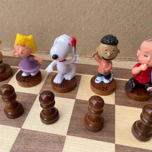 May include: A Peanuts-themed chess set with characters like Charlie Brown, Snoopy, Lucy, and Linus. The chess pieces are made of plastic and are painted in bright colors. The chessboard is made of wood and has a light brown finish.