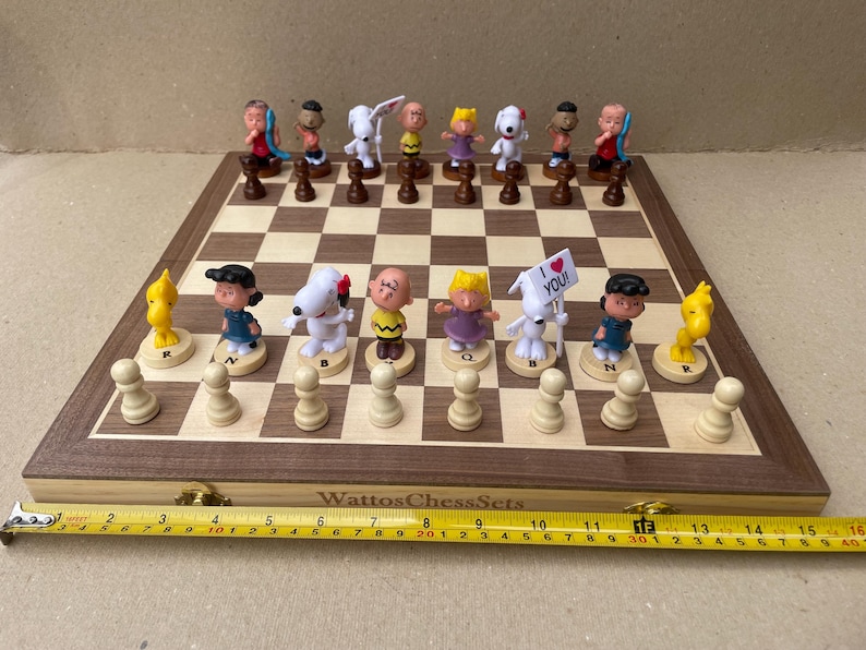 May include: A wooden chessboard with a walnut finish and a set of Peanuts character chess pieces. The chessboard has a light brown and dark brown checkered pattern. The chess pieces are made of plastic and feature characters from the Peanuts comic strip, including Charlie Brown, Snoopy, Lucy, and Linus. The chessboard is set up for a game of chess, with the white pieces on one side and the black pieces on the other. The chessboard is surrounded by a wooden frame.