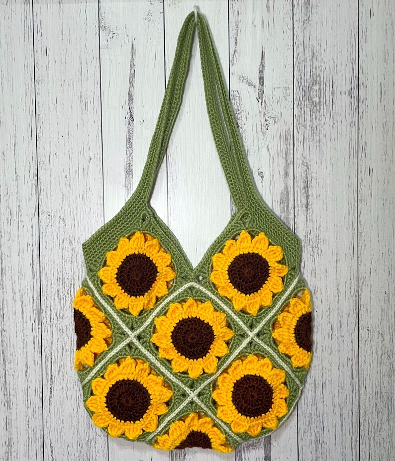 May include: A green crocheted tote bag with a sunflower design. The bag has a square shape and is made of a soft, textured yarn. The sunflowers are yellow with brown centers and are arranged in a repeating pattern.