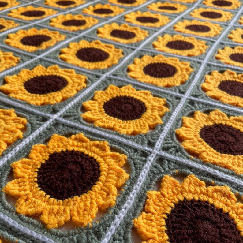 May include: A crocheted blanket featuring a repeating pattern of yellow sunflowers with brown centres. The blanket is made with green and white yarn.