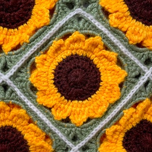 May include: Close-up of a crocheted sunflower blanket with a green and white background. The sunflower is yellow with a brown center.