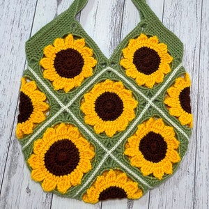 May include: A crocheted tote bag with a green background and a pattern of yellow sunflowers. The bag has two straps and is made with a granny square stitch.