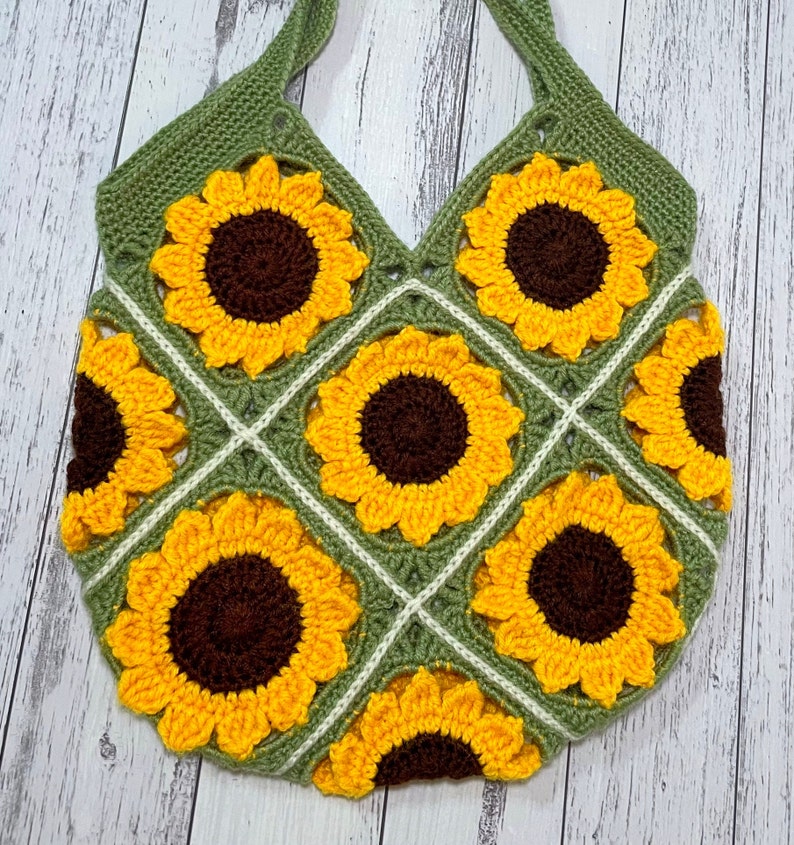 May include: A crocheted tote bag with a green background and a pattern of yellow sunflowers. The bag has two straps and is made with a granny square stitch.