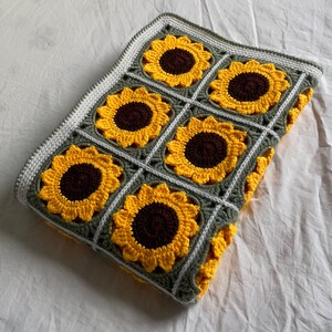 May include: A crocheted blanket featuring a sunflower design in shades of yellow and brown. The blanket is crafted with green and white yarn and has a square shape.