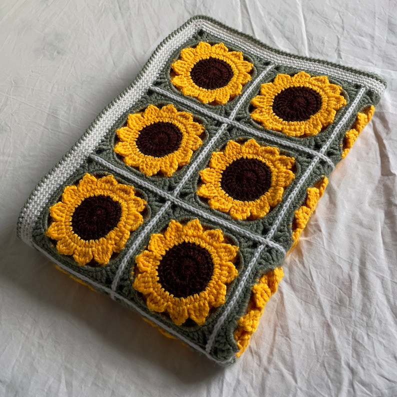 May include: A crocheted blanket featuring a sunflower design in shades of yellow and brown. The blanket is crafted with green and white yarn and has a square shape.