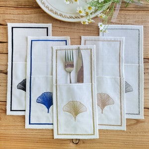 May include: Five white linen placemats with embroidered ginkgo leaf designs in black, blue, gold, brown, and grey. Each placemat has a pocket for holding cutlery.