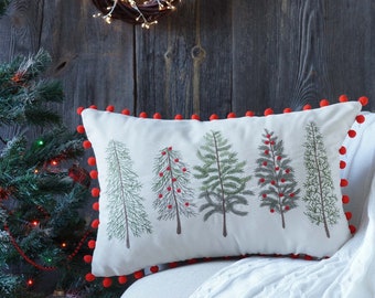 Puede incluir: Una almohada blanca con ribete de pompones rojos presenta un diseño bordado de cinco árboles de Navidad verdes con bayas rojas. La almohada está sentada sobre una manta de punto blanco en un sillón blanco.