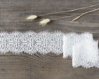 May include: White lace trim with a scalloped edge, displayed on a rustic wooden surface. The lace is intricately patterned and rolled up, with a delicate floral design. Dried ornamental grass adds a touch of natural elegance.