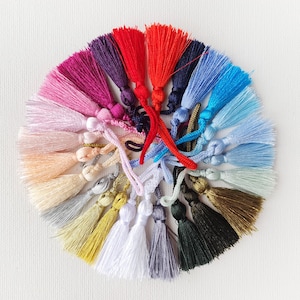 Könnte beinhalten: Eine Sammlung bunter Quasten, die kreisförmig angeordnet sind. Die Quasten sind in verschiedenen Farbtönen gehalten, darunter Rot, Rosa, Lila, Blau, Grün, Gelb und Weiß. Jede Quaste hat eine Schlaufe oben und einen langen, gefransten Boden. Der Hintergrund ist schlicht weiß.