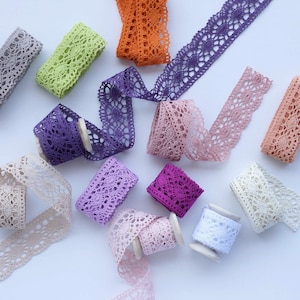 May include: Assortment of colourful lace trim in various shades of purple, pink, beige, green, and orange. The lace trim is arranged on a white background.