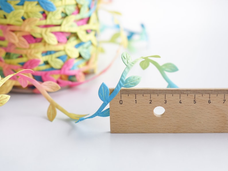 May include: A close-up of a colorful, leafy garland with a wooden ruler measuring 7 inches. The garland is made of paper and features a gradient of blue, green, and yellow leaves.