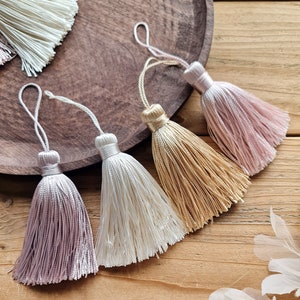 Peut inclure: Cinq petits pompons dans des tons de rose, crème et or. Les pompons sont faits d'un tissu doux et soyeux et ont une frange délicate.