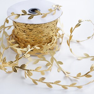May include: A spool of gold leaf ribbon with a leaf design. The ribbon is wound around a white spool and is unfurling to the right.