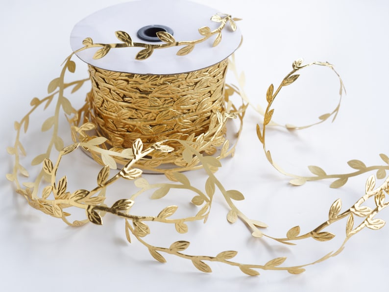 May include: A spool of gold leaf ribbon with a leaf design. The ribbon is wound around a white spool and is unfurling to the right.