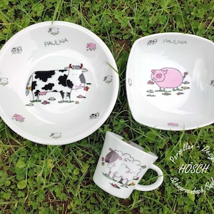 Könnte beinhalten: Ein weißes Kindergeschirr-Set aus Keramik mit einem Kuh-, Schwein- und Schaf-Design. Das Set beinhaltet eine Schüssel, einen quadratischen Teller und eine Tasse. Der Name "PAULINA" ist auf jedem Stück geschrieben.
