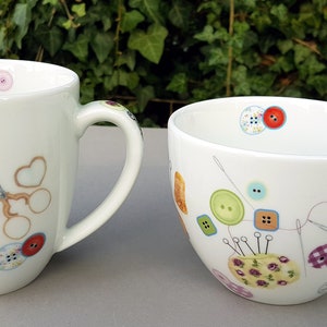 Könnte beinhalten: Zwei weiße Keramikbecher mit bunten Knopf- und Nähmotiven. Der größere Becher hat einen Henkel auf der rechten Seite und der kleinere Becher einen Henkel auf der linken Seite.