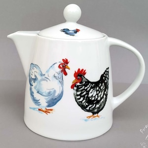 Könnte beinhalten: Weiße Keramik-Teekanne mit Deckel und Henkel. Die Teekanne ist mit Aquarell-Illustrationen von Hühnern in Blau, Schwarz und Weiß verziert. Der Deckel zeigt eine kleine Hühnerillustration. Die Teekanne hat einen Ausguss.