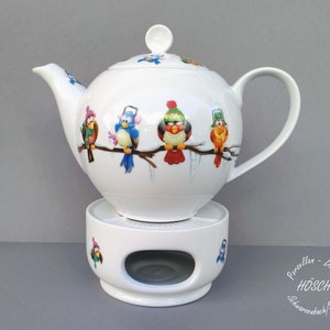 Könnte beinhalten: Weiße Keramik-Teekanne mit passendem Wärmer. Die Teekanne hat ein skurriles Design mit bunten Vögeln, die auf einem Ast sitzen und Wintermützen tragen. Der Wärmer hat eine kreisförmige Öffnung für ein Teelicht.
