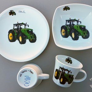 Könnte beinhalten: Ein weißes Porzellan-Kindergeschirr-Set mit grünem Traktor-Design. Das Set beinhaltet einen großen Teller, eine kleine Schüssel und eine Tasse. Der Teller trägt den Namen "PAUL". Die Tasse trägt den Text "von Mama + Papa" auf der Unterseite.