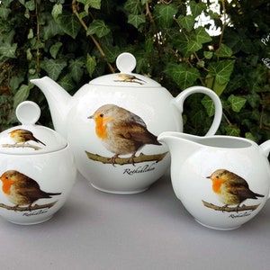 Könnte beinhalten: Ein weißes Porzellan-Tee-Set mit einem Rotkehlchen-Design. Das Set beinhaltet eine Teekanne, eine Milchkännchen und eine Zuckerdose. Jedes Stück zeigt ein Rotkehlchen mit roter Brust und braunen Flügeln, das auf einem Ast sitzt. Der Text "Rotkehlchen" ist auf jedes Stück gedruckt.