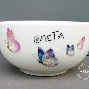 Könnte beinhalten: Weiße Keramikschüssel mit Schmetterlingsdesign. Die Schüssel trägt den Namen "GRETA" in schwarzer Schrift.