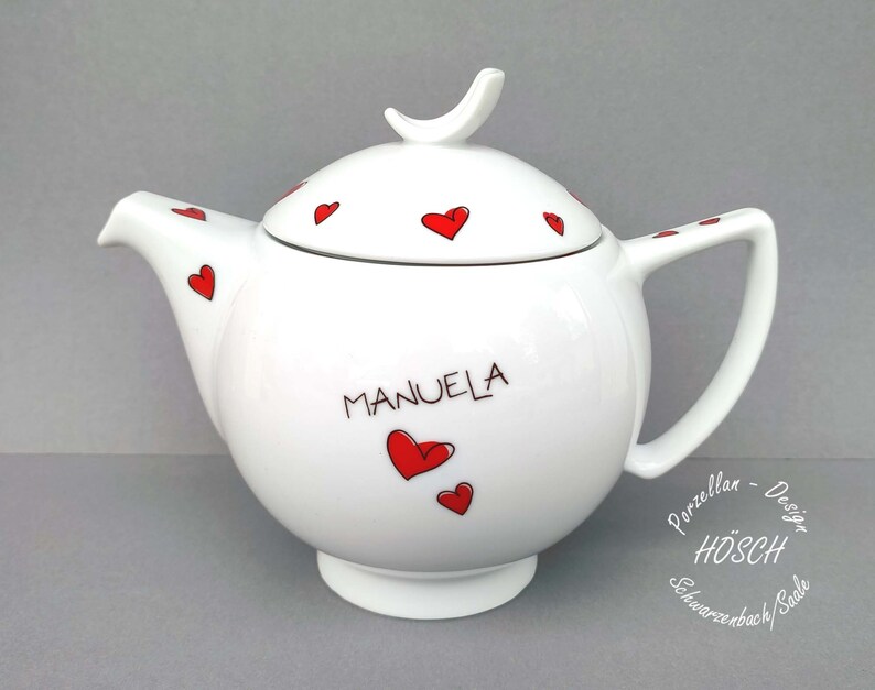 Puede incluir: Tetera de porcelana blanca con un dise&ntilde;o de corazones rojos y el nombre "MANUELA" escrito en el lateral. La tetera tiene una tapa con un asa en forma de p&aacute;jaro y est&aacute; decorada con corazones rojos. La tetera est&aacute; hecha por H&Ouml;SCH Schwarzenbach/Saale.
