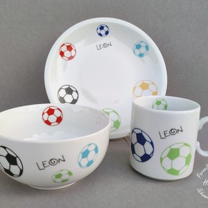 Könnte beinhalten: Ein weißes Kindergeschirr-Set aus Keramik mit Fußball-Motiv. Das Set besteht aus einer Schüssel, einem Teller und einer Tasse. Jedes Stück ist mit einem Fußball-Design und dem Namen "LEON" in schwarzer Schrift verziert.