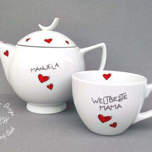 Puede incluir: Conjunto de tetera y taza de t&eacute; blancas con corazones rojos y letras negras. La tetera tiene el nombre "MANUELA" en ella. La taza tiene el texto "WELT BESTE MAMA" en ella.