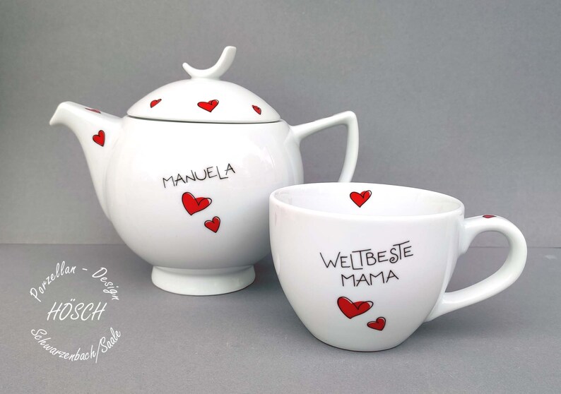Puede incluir: Conjunto de tetera y taza de t&eacute; blancas con corazones rojos y letras negras. La tetera tiene el nombre "MANUELA" en ella. La taza tiene el texto "WELT BESTE MAMA" en ella.
