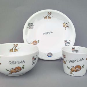 Könnte beinhalten: Ein weißes Kindergeschirr-Set aus Keramik mit einer Schüssel, einem Teller und einem Becher. Jedes Teil ist mit bunten Zeichentierfiguren wie Giraffen, Löwen und Affen verziert. Der Name "ROMINA" ist auf jedem Artikel aufgedruckt. Das Set ist für Kinder konzipiert.