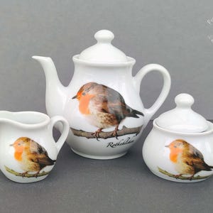Könnte beinhalten: Ein weißes Keramik-Teeservice mit Teekanne, Milchkanne und Zuckerdose. Jedes Teil ist mit einer detaillierten Illustration eines Rotkehlchens mit orange-braunem Gefieder auf einem Ast verziert. Die Teekanne hat einen gebogenen Ausguss und Henkel.