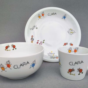 Könnte beinhalten: Ein weißes Kindergeschirr aus Keramik mit einem bunten Comic-Design. Das Set beinhaltet eine Schüssel, einen Teller und eine Tasse. Die Schüssel und die Tasse haben den Namen "Clara" darauf geschrieben. Der Teller hat die Worte "Hallo!" und "Viel Glück" darauf geschrieben.