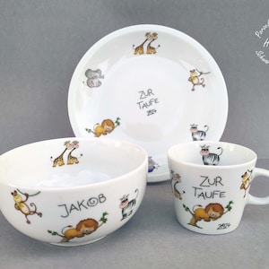 Könnte beinhalten: Ein weißes Keramik-Baby-Set bestehend aus Schüssel, Teller und Tasse mit Dschungeltier-Motiv. Die Schüssel trägt den Namen "Jakob". Teller und Tasse haben beide den Text "Zur Taufe 2024" aufgedruckt.