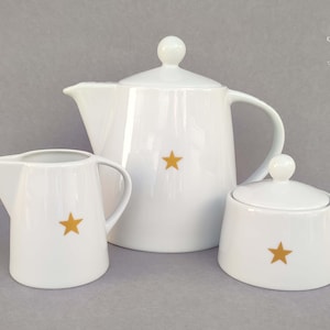 Könnte beinhalten: Weißes Porzellan-Teeservice mit Teekanne, Sahnekännchen und Zuckerdose. Jedes Teil hat ein goldenes Stern-Design. Die Teekanne hat einen Deckel und einen Griff. Das Sahnekännchen hat einen Griff und einen Ausguss.
