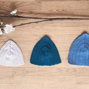 May include: Three crocheted hats in different colours: white, teal, and light blue. The hats are arranged in a row on a wooden surface.