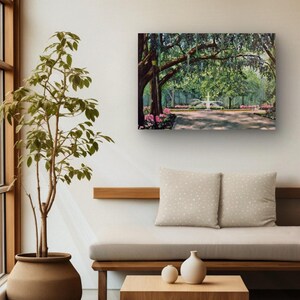 May include: A framed painting depicts a park scene with a fountain, trees, and a pathway. The artwork features lush greenery, pink flowers, and a light-filled atmosphere. The painting hangs on a white wall above a wooden bench with two pillows.
