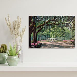 May include: A framed painting depicts a park scene with a fountain, trees, and flowers. The artwork features a pathway leading to a fountain, surrounded by lush greenery and pink flowers. The painting is displayed on a white shelf with decorative plants.