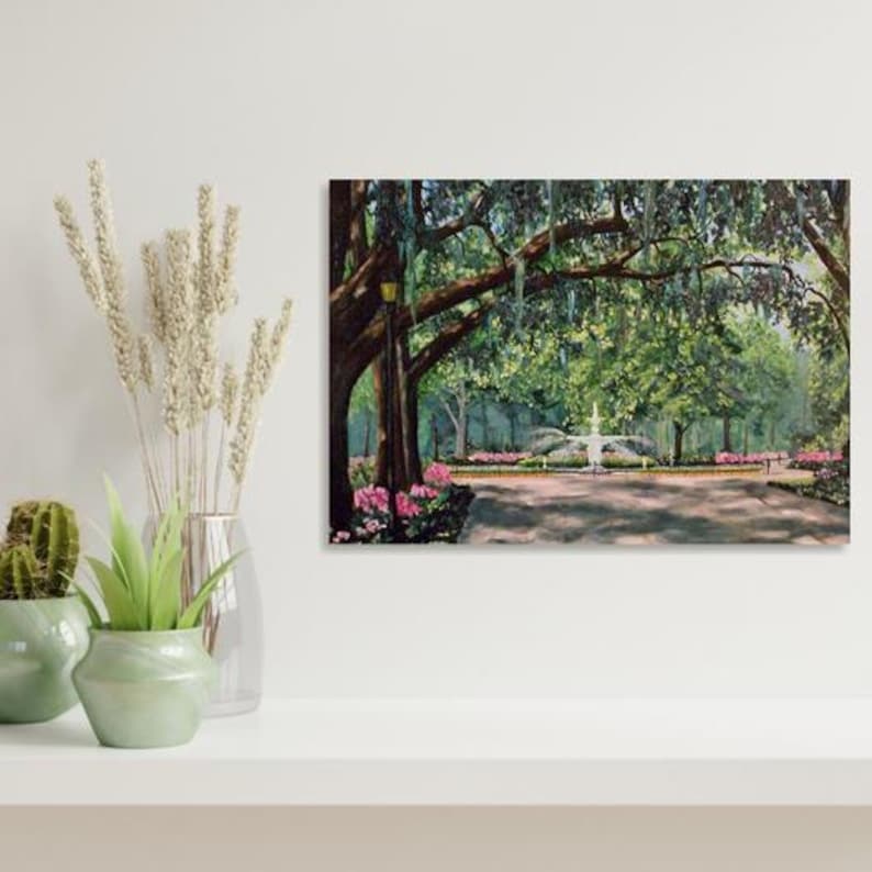 May include: A framed painting depicts a park scene with a fountain, trees, and flowers. The artwork features a pathway leading to a fountain, surrounded by lush greenery and pink flowers. The painting is displayed on a white shelf with decorative plants.