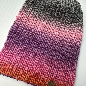 May include: A knitted beanie hat with a colour gradient. The hat fades from black and white at the crown, through pink and purple, to orange at the brim. A small brown leather tag is attached. The hat is made of soft yarn.
