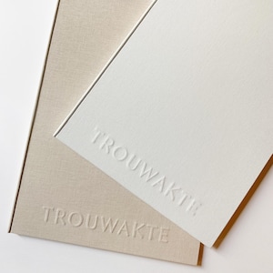 Op de afbeelding: Twee hardcover boeken, één crème en één wit, met het woord "TROUWAKTE" in reliëf op de omslagen. Het crème boek heeft een getextureerde linnen-achtige afwerking. Een witte bloem en een gouden haaraccessoire zijn op de achtergrond.