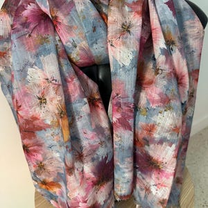 May include: A lightweight scarf featuring a watercolour-style floral print in shades of dusty blue, magenta, soft pink, and hints of orange. The fabric has a visible weave texture and frayed edges, displaying a painterly pattern of scattered blossoms.