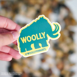 May include: A yellow and teal sticker with a silhouette of a woolly mammoth. The sticker says "WOOLY" in yellow letters and "@WOOLYPENNANTS" in smaller yellow letters at the bottom.