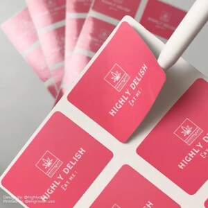 May include: Pink square stickers with a white outline and a white graphic of a marijuana leaf inside a square with the text "HIGHLY DELISH EAT ME!"