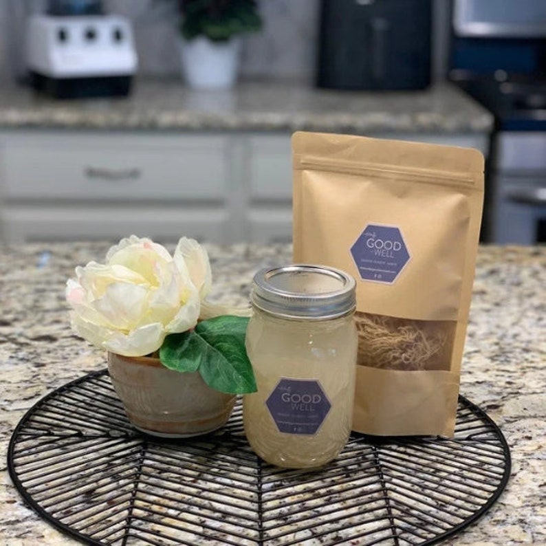 May include: A glass jar filled with clear liquid and a brown paper bag with the text "The Good Well" on it. The jar and bag are sitting on a black metal tray.