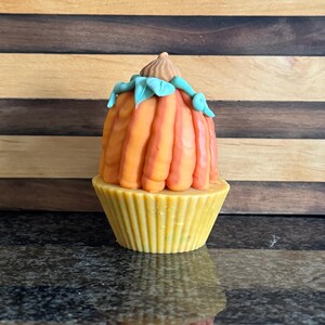 以下が含まれることがあります： 黄色いカップケーキの上に、オレンジと黄色のパンプキンフロスティングと緑の葉が乗っています。