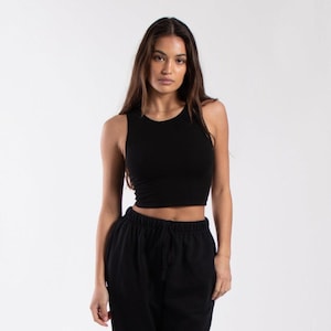 Könnte beinhalten: Ein schwarzes Crop-Top mit rundem Ausschnitt und ärmellosem Design. Das Oberteil wird mit einer schwarzen Jogginghose kombiniert. Das Model steht vor einem weißen Hintergrund.